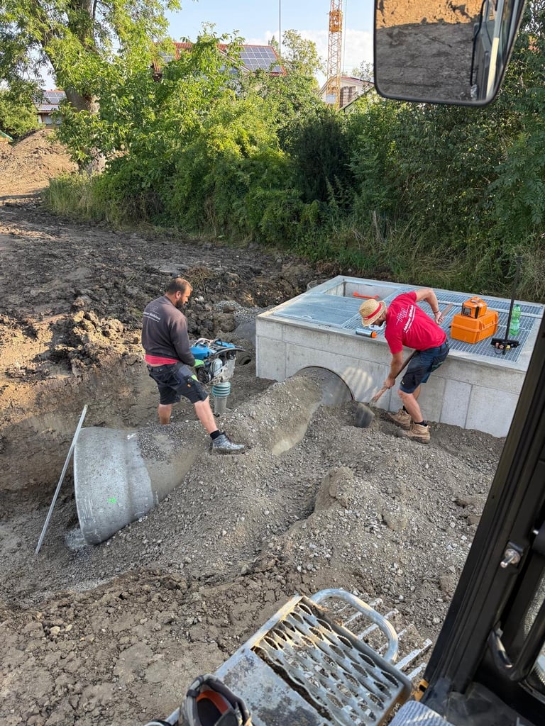 Zwei Bauarbeiter bei der Montage eines großen Betonrohrs auf einer Baustelle mit grüner Vegetation