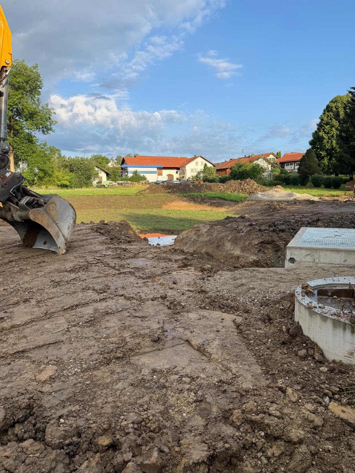 Baustelle mit Baggerlöffel, Betonrohren und Wohnhäusern im Hintergrund unter blauem Himmel