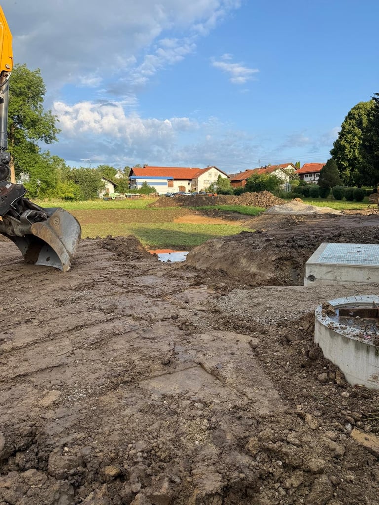 Baustelle mit Baggerlöffel, Betonrohren und Wohnhäusern im Hintergrund unter blauem Himmel