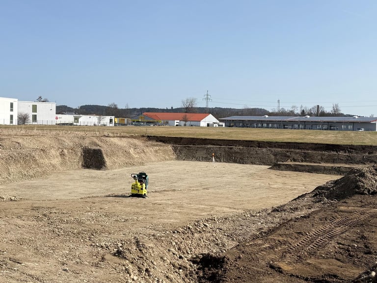Bauarbeiter auf ausgehobenem Grundstück mit Gebäuden und landwirtschaftlichen Strukturen im Hintergrund unter klarem Himmel