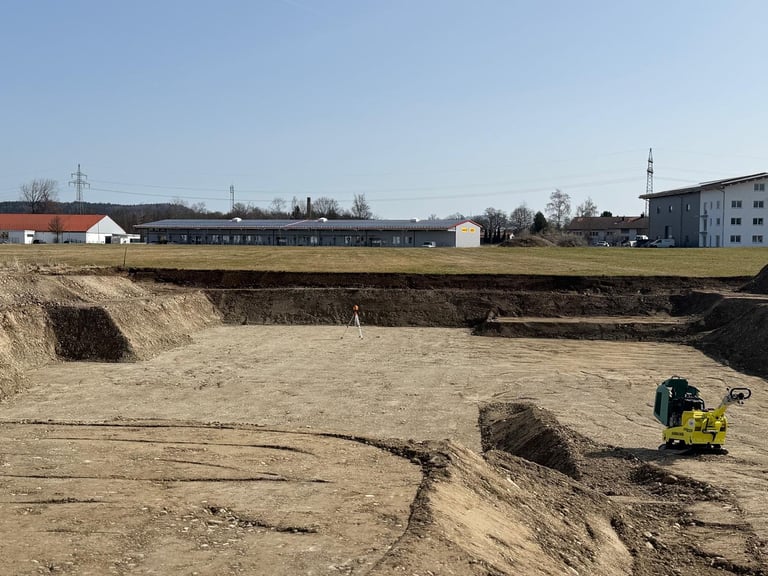Baustelle mit ausgehobener Erde, schweren Maschinen, Ackerland und Wohngebäuden im Hintergrund unter klarem Himmel