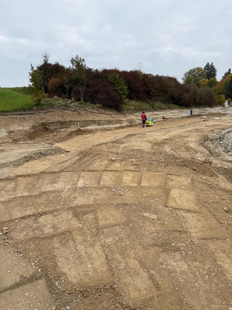 Baustelle mit gerodeter Erdfläche, Arbeiter in Warnweste, Bäumen und grünen Feldern im Hintergrund
