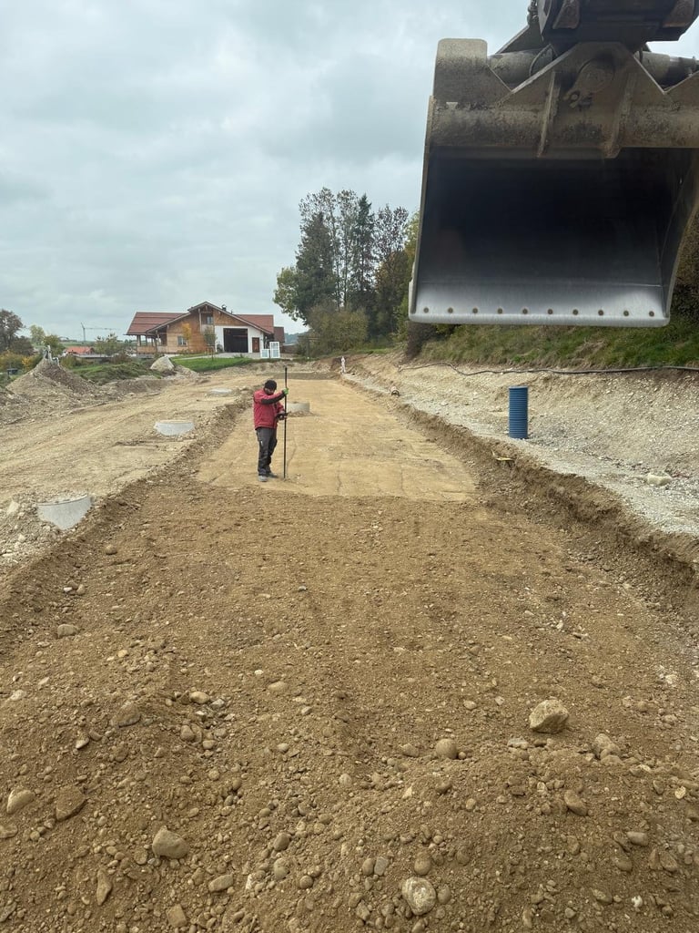 Baustelle mit Arbeiter in roter Jacke beim Vermessen des Erdreichs, großer Baggerlöffel sichtbar, ländliche Gebäude im Hintergrund