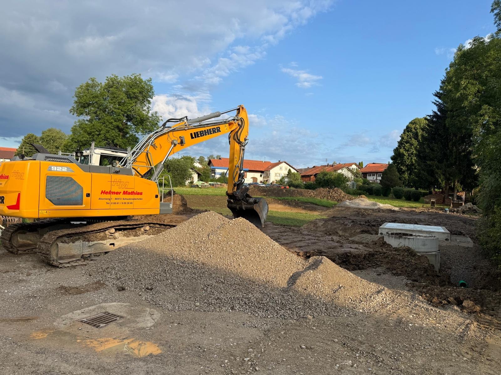 Gelber Bagger lädt Kies auf einer Baustelle mit Wohnhäusern im Hintergrund