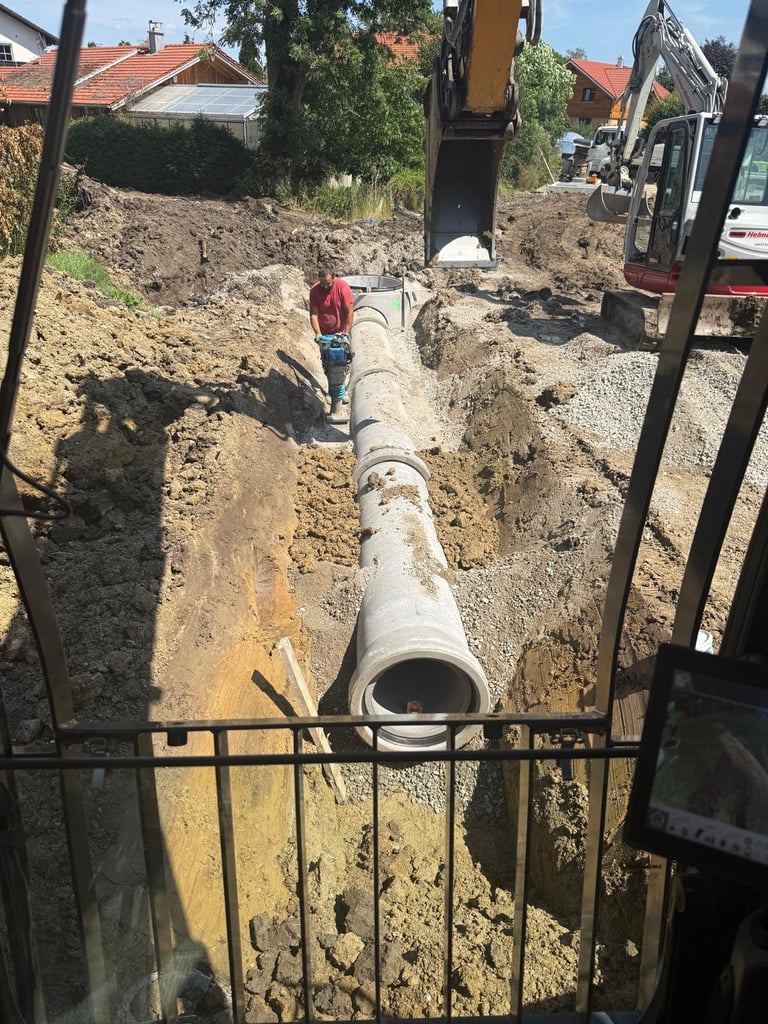Großes Betonabflussrohr in ausgehobenem Graben auf einer Baustelle mit Bagger und Arbeiter