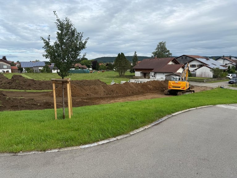 Baustelle mit gelbem Bagger beim Bewegen von Erde, neu gepflanzter Baum mit Stützpfählen, asphaltierte Straße, grünes Gras und Wohnhäuser im Hintergrund unter bewölktem Himmel