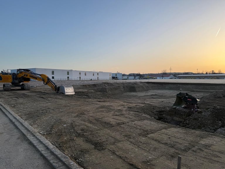 Baustelle bei Sonnenuntergang mit Bagger, Erdhaufen und Industriegebäuden im Hintergrund unter klarem Himmel