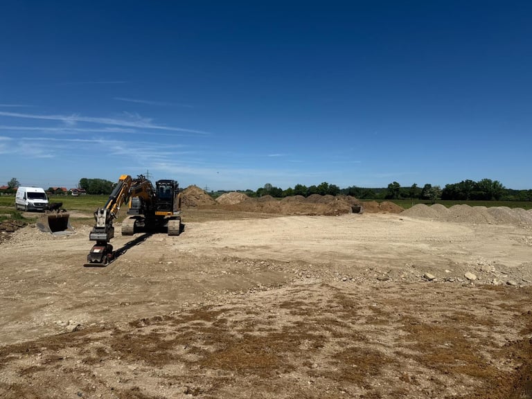 Bagger und weißer LKW auf einer Baustelle mit gerodetem Sandboden und Bäumen im Hintergrund unter blauem Himmel
