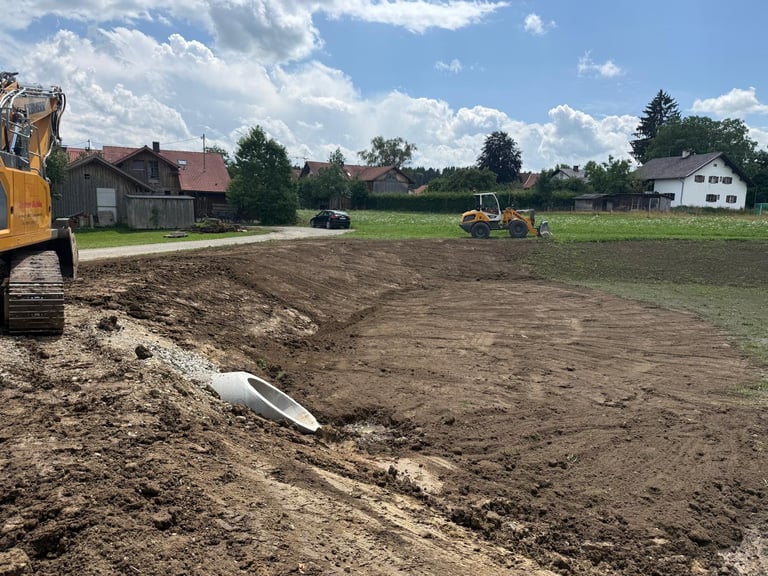 Baustelle mit gelbem Bagger und Grader auf braunem Erdreich neben ländlichen Gebäuden