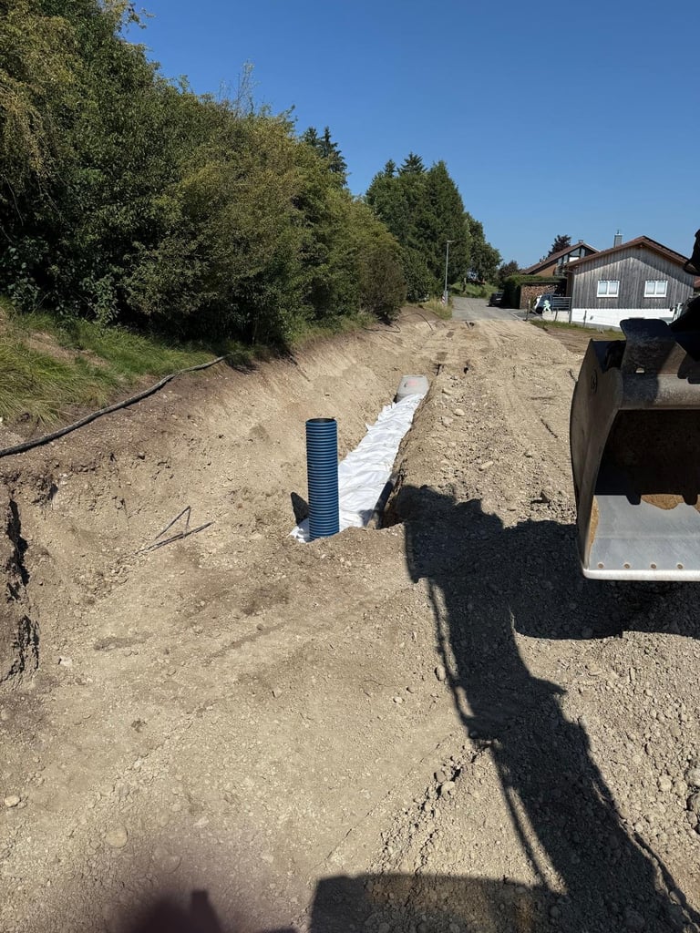 Baustelle mit blauem Entwässerungsrohr auf sandigem Boden, weißem Vlies, Bäumen und Wohngebäude unter klarem Himmel